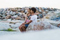 Joven abrazando pareja sentada en rocas en la costa y mirando hacia otro lado . - foto de stock