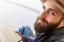 Bearded man listening to music with book in hands and looking at camera — Stock Photo