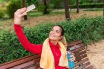 Спортивна дівчина з рушником на плечах і пляшкою води, що сидить на лавці парку і приймає селфі — стокове фото