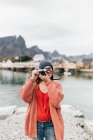 Woman in knitted cardigan and hat taking pictures on analog camera at mountain lake. — Stock Photo