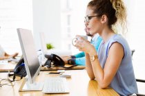 Vista lateral da mulher sentada no local de trabalho com copo e olhando para o monitor — Fotografia de Stock