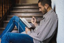Вид сбоку на юного студента в клетчатой рубашке, сидящего на лестнице и пишущего в блокноте . — стоковое фото