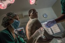 BENIN, ÁFRICA - 31 de agosto de 2017: Hombre negro sentado en el hospital y mirando por encima del hombro a la cámara mientras el personal de tratamiento . - foto de stock