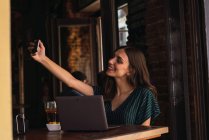 Porträt einer Frau, die mit Laptop am Cafétisch sitzt und ein Selfie macht — Stockfoto