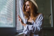 Redhead woman in hat posing at window with shutters — Stock Photo