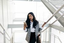 Elegante mujer de negocios subir escaleras y mirando el teléfono inteligente en la mano - foto de stock