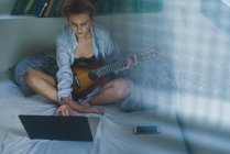 Low angle view of girl sitting on bed with guitar and using laptop — Stock Photo