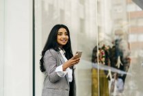 Портрет веселої бізнес-леді, що тримає смартфон і дивиться в сторону вікна магазину — стокове фото