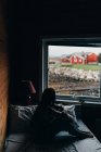 Vista lateral da silhueta feminina sentada na cama no quarto escuro e olhando para casas vermelhas atrás da janela . — Fotografia de Stock