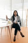 Элегантная деловая женщина со смартфоном сидит за столом с чашкой чая и смотрит в камеру — стоковое фото