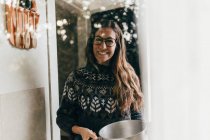 Cheerful brunette woman in glasses holding pot and looking at camera through window — Stock Photo