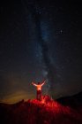 Rückansicht eines rotbeleuchteten Mannes, der mit erhobenen Armen unter Milchstraße im Himmel posiert — Stockfoto