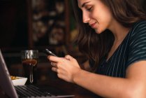 Donna sorridente che chiacchiera sullo smartphone mentre è seduta al computer portatile in un caffè . — Foto stock