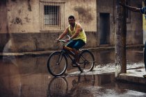 CUBA - AUGUST 27, 2016: Side view of man riding bicycle through paddle on background of poor city district. — Stock Photo