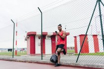 Sportsman dans les écouteurs penché sur la clôture et smartphone de navigation — Photo de stock