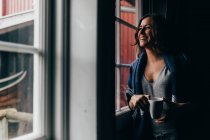 Усміхнена жінка з чашкою дивиться у вікно — стокове фото