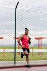 Спортсмен в наушниках, опирающийся на забор и просматривающий смартфон после тренировки — стоковое фото
