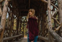 Chica morena en chaqueta con capucha roja caminando en pasaje de madera y mirando por encima del hombro a la cámara - foto de stock