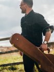 Vista lateral do homem vestindo camisa jeans posando com caixa de guitarra em campo — Fotografia de Stock
