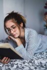 Portrait of brunette girl in glasses reading book and lying on bed — Stock Photo