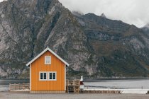 Small cabin standing on lake shore on background of massive mountains cliffs. — Stock Photo