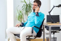Retrato de un joven hombre de negocios sentado en silla y hablando por teléfono inteligente en la oficina moderna . - foto de stock