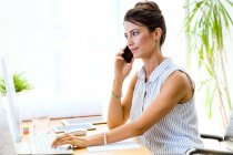 Portrait of businesswoman using laptop and talking on smartphone in modern office. — Stock Photo
