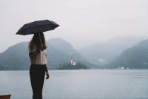 Femme avec parapluie posant sur la rive du lac et regardant loin — Photo de stock