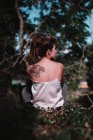 Вид сзади девушки с татуировкой на спине, сидящей на заборе в саду — стоковое фото
