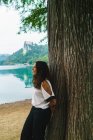 Seitenansicht eines brünetten Mädchens, das sich am Ufer des Sees an einen Baumstamm lehnt — Stockfoto