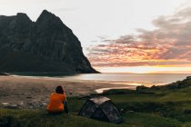 Вид спереду жінки, що сидить на пагорбі біля намету і дивиться на світанковий морський пейзаж — стокове фото