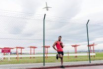 Esportista em fones de ouvido apoiados na cerca com smartphone na mão e olhando para longe — Fotografia de Stock