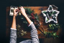 Vista superior de manos femeninas confeccionando corona de Navidad en mesa de madera - foto de stock