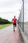 Front view of man wearing headphones running on track along fence — Stock Photo