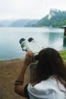 Vista trasera de la mujer morena mirando la máquina binocular de turismo en la orilla del lago - foto de stock