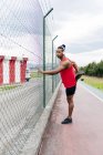 Вид сбоку спортсмена в наушниках, опирающегося на проволочный забор и растягивающего ногу перед тренировкой — стоковое фото