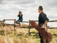 Жінка з старовинною камерою сидить на сільському паркані і знімає чоловіка з гітарою — стокове фото