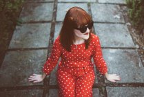 Рыжеволосая женщина в ярко-красном костюме и солнечных очках, счастливо позирующая на земле . — стоковое фото
