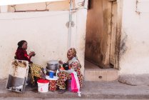 Goree, Sénégal- 6 décembre 2017 : Vue de côté de la femme assise dans la rue près de la maison nourrissant bébé et parlant à un ami — Photo de stock