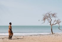 Goree, Senegal- December 6, 2017: Side view of woman in traditional clothes walking on beach near waving sea and tree. — Stock Photo