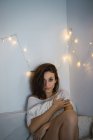 Bruna ragazza seduta sul letto sopra il muro con le luci delle fate e guardando la fotocamera — Foto stock