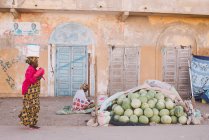 Goree, Senegal- 6 de diciembre de 2017: Vista lateral de la mujer vestida con ropa tradicional llevando cubo en la cabeza y caminando cerca de un montón de sandías . - foto de stock
