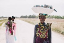 Goree, Senegal- 6 de diciembre de 2017: Retrato de una mujer africana sonriente llevando una cesta en la cabeza y caminando por la carretera . - foto de stock