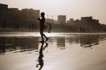 Yoff, Senegal- Dezembro 6, 2017: Silhueta da criança refletindo na superfície da água rasa no fundo da cena de rua na névoa da luz solar
. — Fotografia de Stock