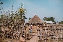 Vista trasera del hombre caminando a bungalows con techo de paja en pueblo africano nativo
. - foto de stock
