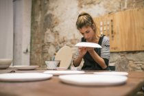 Mujer concentrada en delantal sentada a la mesa y creando platos de arcilla blanca . - foto de stock