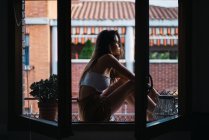 Vista lateral de chica morena relajándose en el alféizar de la ventana en la terraza del apartamento - foto de stock