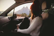 Vista lateral da mulher de chapéu vermelho sentado no carro e olhando para a janela durante a viagem ao campo . — Fotografia de Stock