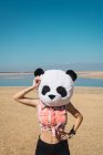 Girl posing in toy head of panda on background of sandy lake beach. — Stock Photo