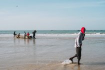 Goree, Senegal- 6 de dezembro de 2017: Vista lateral do homem andando na praia no fundo das pessoas que puxam o barco para o oceano . — Fotografia de Stock
